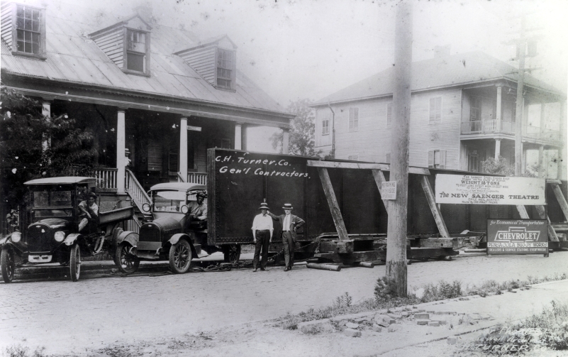An aged black and white photo of historic Pensacola, featuring a large beam on the street with two men posing in front of it. The words 