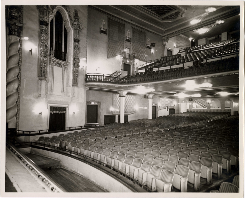 An old, black and white tintype photo of the empty interior of the Saenger Theatre in a Spanish renaissance architectural style with rows of velvet upholstered theatre seats.