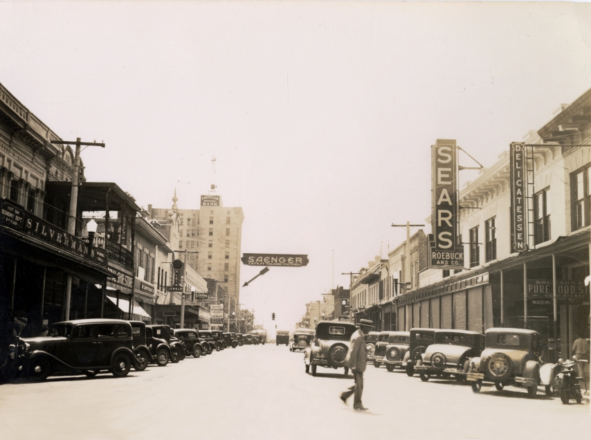An old black and white image of historic Palafox street in the 1930s lined with vintage cars and featuring a large sign pointing to the Saenger Theatre.